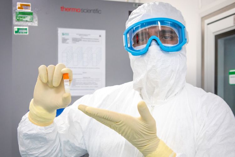 Pessoa paramentada com óculos e roupa especial apontando frasco com cepa do vírus influenza em ambiente de laboratório