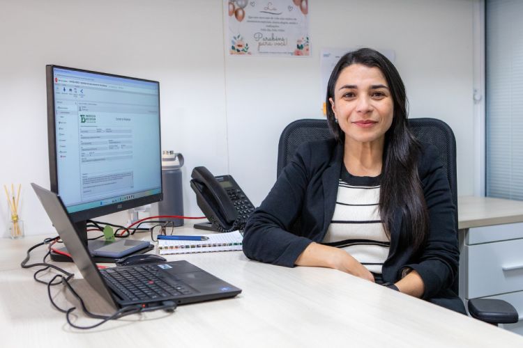 Mulher branca com cabelos longos escuros, sentada em uma mesa com computador