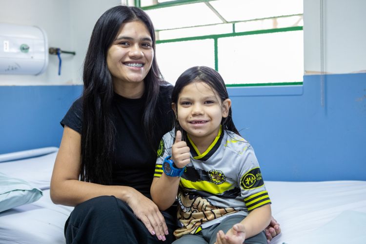 Mulher de cabelos compridos pretos e criança, sentados, em ambiente hospitalar