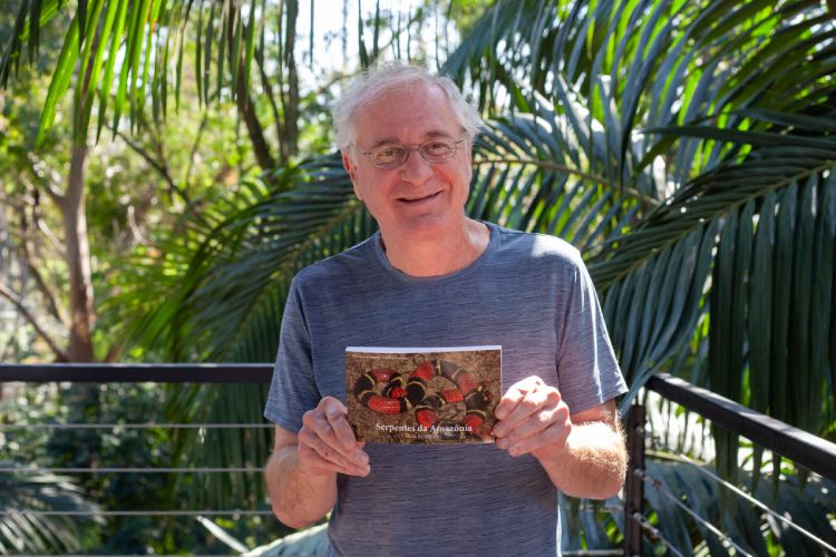 Otavio Marques: homem branco, de cabelos grisalhos e óculos, segurando livro, segurando livro com árvores ao fundo